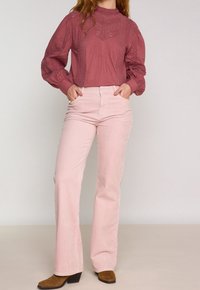 Kvinna klädd i en vinröd spetsblus, ljusrosa kordbyxor och bruna klackade stövlar, stående med händerna i fickorna.