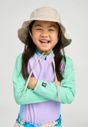 Smiling young girl wearing a beige sun hat, pastel purple and mint green long-sleeve top, and colorful shorts, arms crossed confidently.