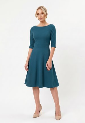 Mujer de pie con un vestido azul verdoso hasta la rodilla con mangas tres cuartos y tacones altos beige sobre un fondo blanco liso.