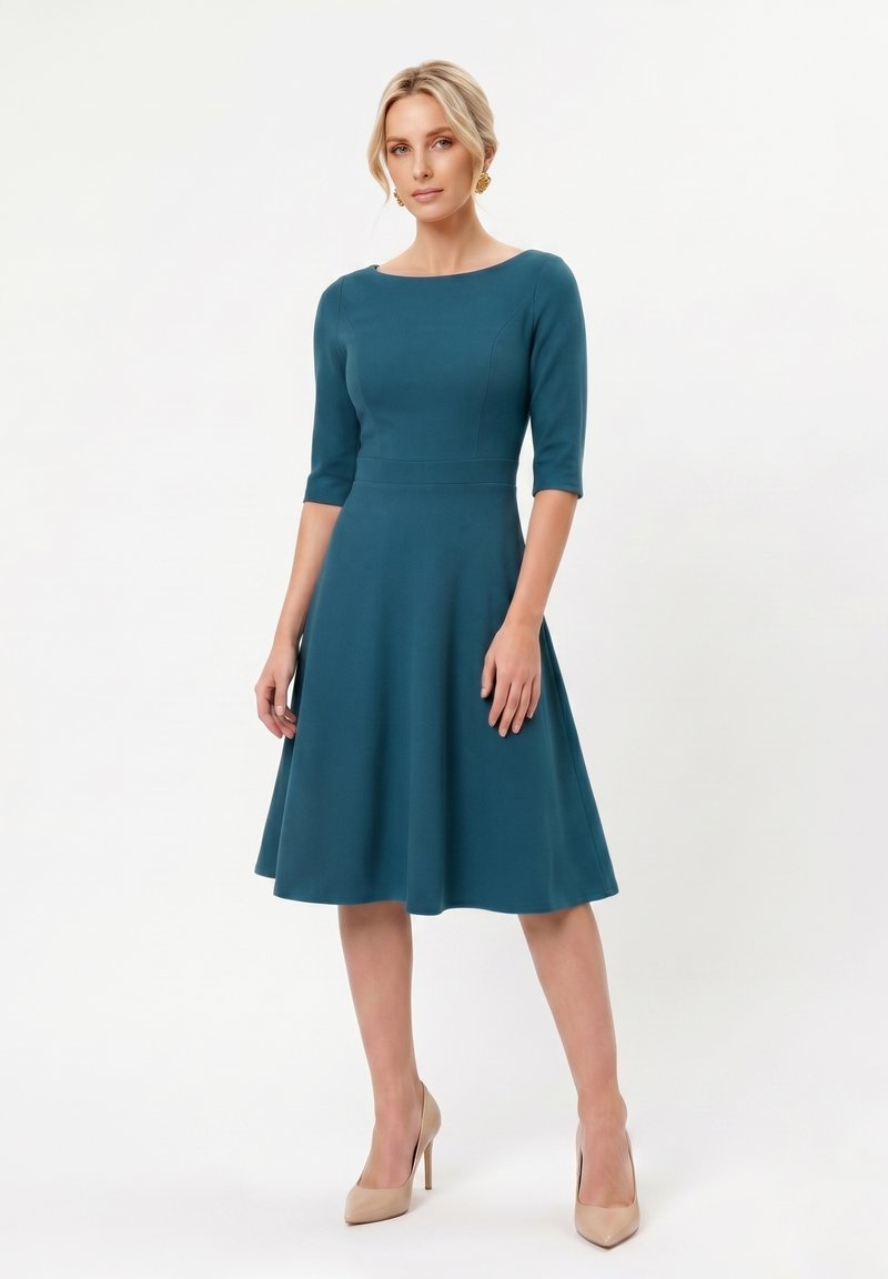 Femme debout portant une robe bleu sarcelle longueur genou avec des manches trois-quarts et des talons hauts beiges sur un fond blanc uni.