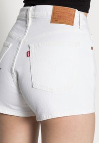 Shorts de mezclilla blanca con una cintura alta, diseñados con bolsillos traseros y una etiqueta de marca de cuero marrón. Presentan costuras clásicas y botones de cobre.