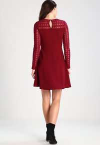 Robe bourgogne avec un corsage ajusté, des manches longues en dentelle et un détail dos en forme de trou de serrure, dotée d'une jupe évasée et d'une texture douce.