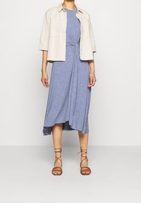Personne portant une veste ouverte beige clair sur une robe midi bleue nouée avec un ourlet asymétrique et des sandales à brides marron.