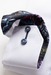 Poignet de chemise blanche avec deux boutons foncés et une doublure intérieure colorée à motifs, replié sur un fond uni.