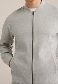 Šedá bomber bunda s herringbonovým vzorem, zapínáním na zip a žebrovanými manžetami. Obsahuje dvě boční kapsy a jemný textilní povrch.