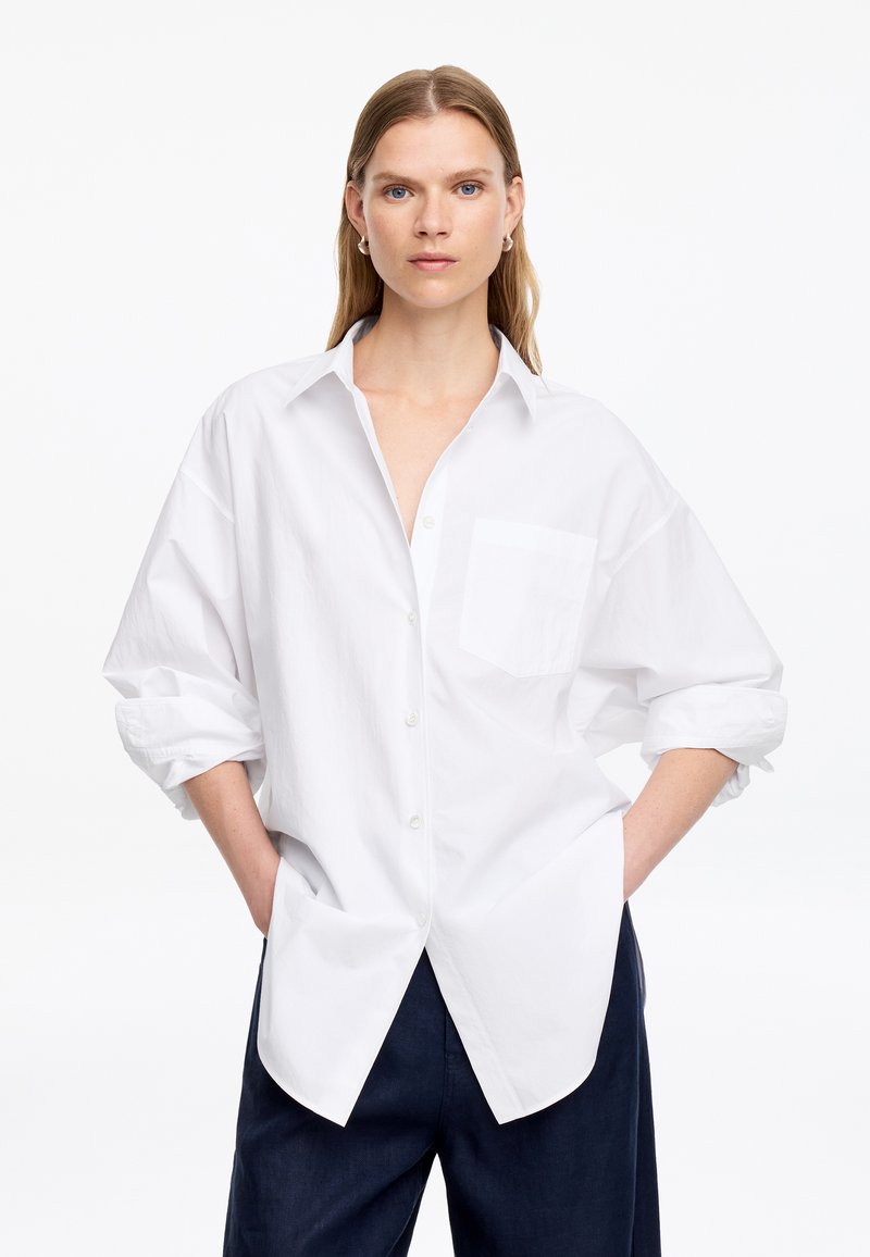 Femme aux longs cheveux blonds porte une chemise blanche ample à boutons avec les manches retroussées et un pantalon foncé, debout les mains dans les poches.