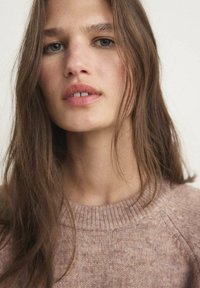 Maglione lavorato a maglia rosa chiaro con colletto rotondo, caratterizzato da un motivo testurizzato e polsini a coste. Il modello presenta un trucco naturale e capelli lunghi.