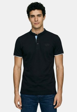 Jeune homme aux cheveux bouclés et foncés portant un polo noir à manches courtes avec bordure blanche et un jean bleu foncé, debout devant un fond uni.