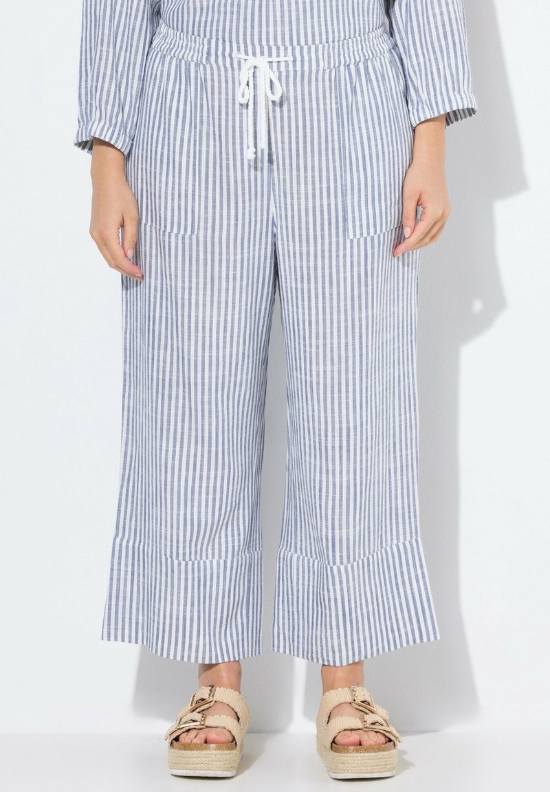 Personne portant un pantalon large à rayures verticales bleu et blanc avec poches avant et des sandales beiges à plateforme, debout sur un fond blanc.