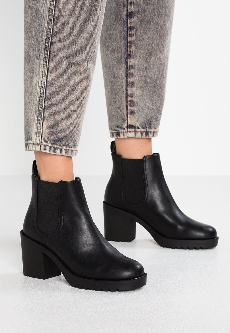 Bottes basses en cuir noir à talons carrés portées avec un jean court gris clair délavé à l'acide, posées sur un sol blanc.