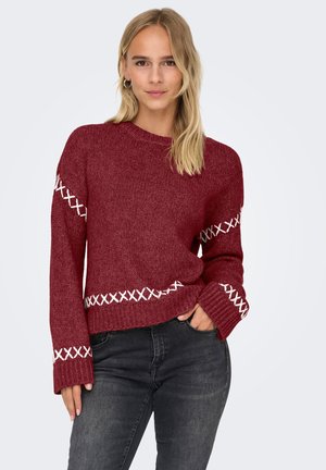 Kvinne med blondt hår iført en rød strikkegenser med hvite broderidetaljer og mørkegrå jeans, poserer med den ene hånden i lomma.