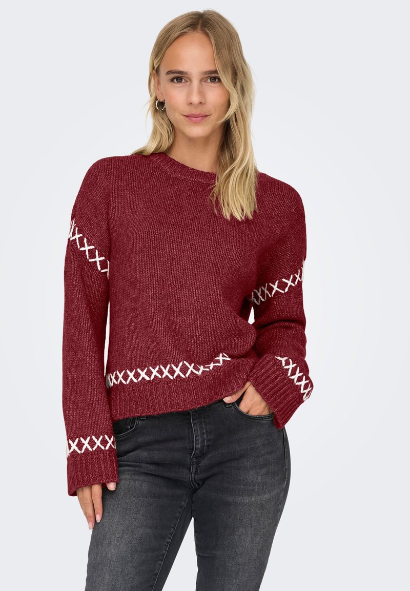 Femme aux cheveux blonds portant un pull en tricot rouge avec des détails en broderie blanche et un jean gris foncé, posant avec une main dans la poche.