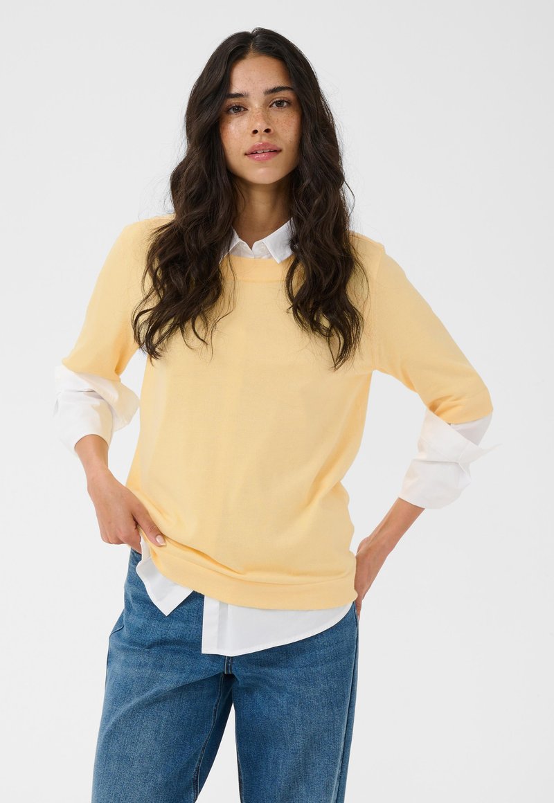 Jeune femme aux longs cheveux foncés portant un pull jaune clair par-dessus une chemise blanche et un jean bleu, debout avec les mains sur les hanches devant un fond blanc.