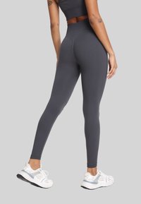 Kobieta w ciemnoszarych legginsach z wysokim stanem i białych sneakersach, pokazana z tyłu na jednolitym szarym tle.