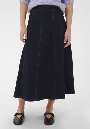 Femme portant une jupe midi en denim bleu foncé, taille haute, coupe trapèze, un pull lavande et des chaussures plates noires avec des brides froncées.