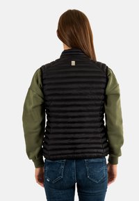 Chaleco acolchado negro con patrón de acolchado horizontal, cuello con cremallera y detalle de logo. Usado sobre una camiseta de manga larga verde, combinado con jeans azules.