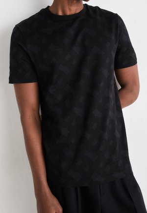 Mand iført en sort, kortærmet t-shirt med et diskret geometrisk mønster mod en ensartet hvid baggrund.