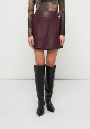 Burgundin nahkainen mini-hame, jossa on kaksi hopeista solkea, yhdistettynä korkeisiin mustiin kärkitossuihin. Tasainen pinta ja tiukka muotoilu.