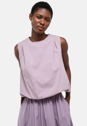 Femme aux cheveux courts portant un haut lavande sans manches et ample, avec une jupe assortie froncée, debout les mains dans les poches.