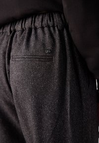 Gros plan sur un pantalon gris foncé texturé avec une ceinture élastique et un petit logo métallique rond au-dessus d'une poche arrière fendue.