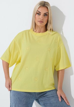 Kobieta o blond włosach, ubrana w luźną żółtą koszulkę z subtelnymi haftowanymi kwiatami, w połączeniu z niebieskimi dżinsami, stojąca na tle białej ściany.