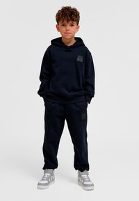 Mörkblå hoodie och matchande mjukisbyxor i mjukt tyg. Har en logotypmärke framtill. Bär grå sneakers med vita detaljer.