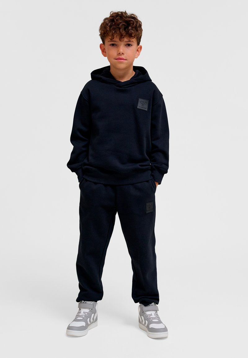 Mörkblå hoodie och matchande mjukisbyxor i mjukt tyg. Har en logotypmärke framtill. Bär grå sneakers med vita detaljer.