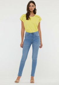 Haut jaune à manches courtes avec des accents en dentelle, associé à un jean skinny bleu clair et des sandales beiges. Design simple, texture lisse, coupe ajustée.