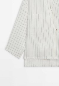 Veste blanche à fines rayures verticales noires, fermeture à un bouton, manches longues, et pantalon assorti à rayures partiellement visible en dessous.