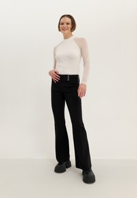 White long-sleeve top with sheer accents, paired with black flared trousers featuring a zip and button closure, worn with chunky black shoes.