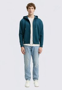 Türkiz zipzáras kapucnis pulóver húzózsinórokkal, fehér ing fölött viselve. Világoskék farmerral és fehér tornacipővel párosítva, laza megjelenést kölcsönözve.