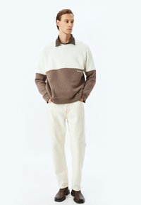 Jersey en color crema y marrón, diseño en bloques de color, cuello redondo, puños acanalados. Pantalones crema y botas marrones oscuras completan el conjunto.