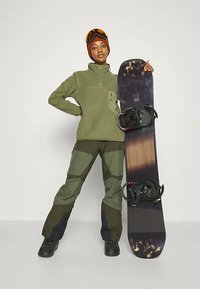 Snowboarder i en olivengrøn fleecejakke, grønt og sort mønstrede bukser, sorte støvler, holder en mørk snowoard med bindinger.