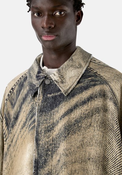 Jeune homme portant une veste beige et noire à motif avec un col, par-dessus un pull côtelé de couleur claire, regardant droit devant.