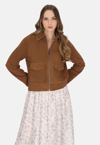 Giacca cropped in suede marrone con zip frontale, due tasche anteriori e colletto. Indossata sopra una gonna bianca con motivo floreale.