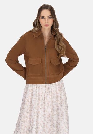 Bruine suède cropped jack met een ritssluiting aan de voorkant, twee voorzakken en een kraag. Draag hem over een witte rok met bloemenpatroon.
