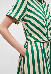 Πράσινο και μπεζ φόρεμα με ρίγες, κοντά μανίκια, κουμπιά στο μπροστινό μέρος και ζώνη στη μέση. Διαθέτει πλαϊνή τσέπη και ομαλή υφή υφάσματος.