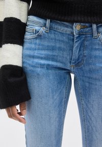 Ανοιχτό μπλε τζιν με ελαφρώς φθαρμένη υφή, με χάλκινο κουμπί, πέντε τσέπες και εφαρμοστή γραμμή.