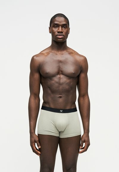 Μυώδης άνδρας, όρθιος και στραμμένος προς τα εμπρός, που φοράει ανοιχτό γκρι κοντά εσώρουχα boxer με μαύρη μέση, πάνω σε απλό λευκό φόντο.