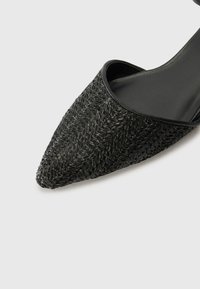 Zwarte puntige schoen met geweven textuur op de bovenkant, voorzien van een gladde zwarte leren afwerking en een lage, gebogen hak.