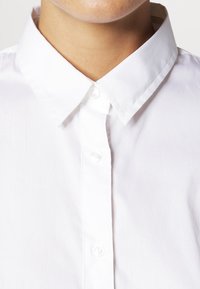 Primo piano di una persona che indossa una camicia bianca a bottoni con colletto a punta su uno sfondo neutro.