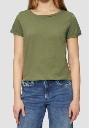 Młoda kobieta ubrana w oliwkowo-zielony t-shirt z krótkim rękawem i niebieskie dżinsy stoi na neutralnym tle.