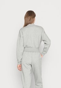 Person med långt brunt hår som bär en grå sweatshirt och matchande träningsbyxor, står med en hand i fickan, vänd bort.