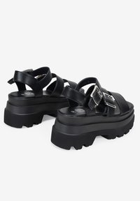 Sandali platform neri con suola spessa, cinturini incrociati e dettaglio in metallo sulla fibbia. Il materiale è liscio con una finitura testurizzata.