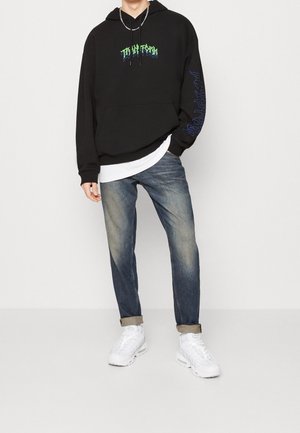 Man draagt een zwarte hoodie met groene tekst, vervaagde blauwe jeans opgerold bij de enkels, witte sneakers en een wit shirt eronder.