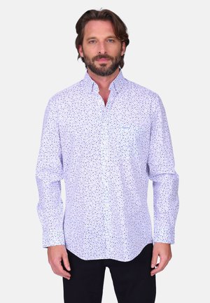Homme barbu portant une chemise blanche à manches longues avec de petits motifs floraux bleus, debout devant un fond uni.
