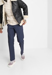 Schwarze Lederjacke mit weißem Schaffellfutter, beige Kapuzenpullover, marineblaue Hose und gemusterte Sneaker in Weiß und Burgunderrot.