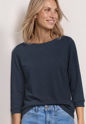 Blond vrouw die lacht, gekleed in een donkerblauw shirt met lange mouwen en lichtblauwe spijkerbroek, licht naar rechts gekeerd.