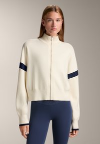 Cremfarbene Zip-Jacke mit hohem Kragen, verziert mit marineblauen Streifenakzenten an den Ärmeln. Weicher Stoff, lockere Passform und verkürzte Silhouette.