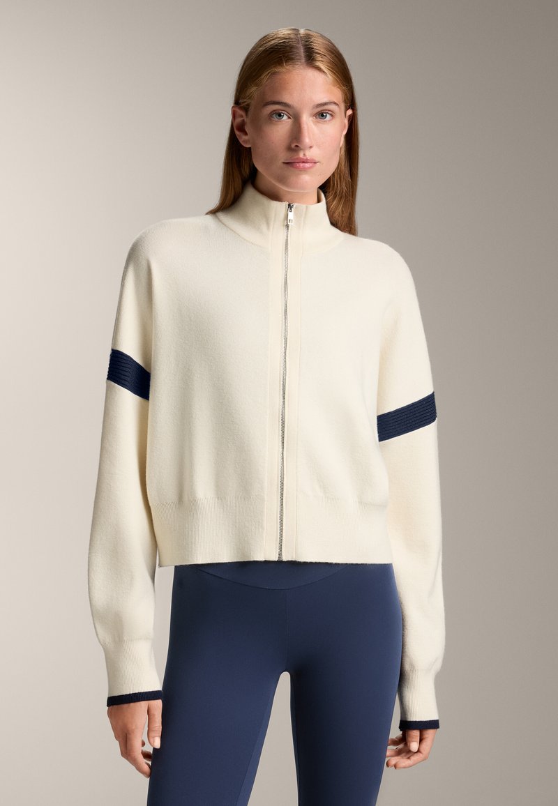 Cremfarbene Zip-Jacke mit hohem Kragen, verziert mit marineblauen Streifenakzenten an den Ärmeln. Weicher Stoff, lockere Passform und verkürzte Silhouette.